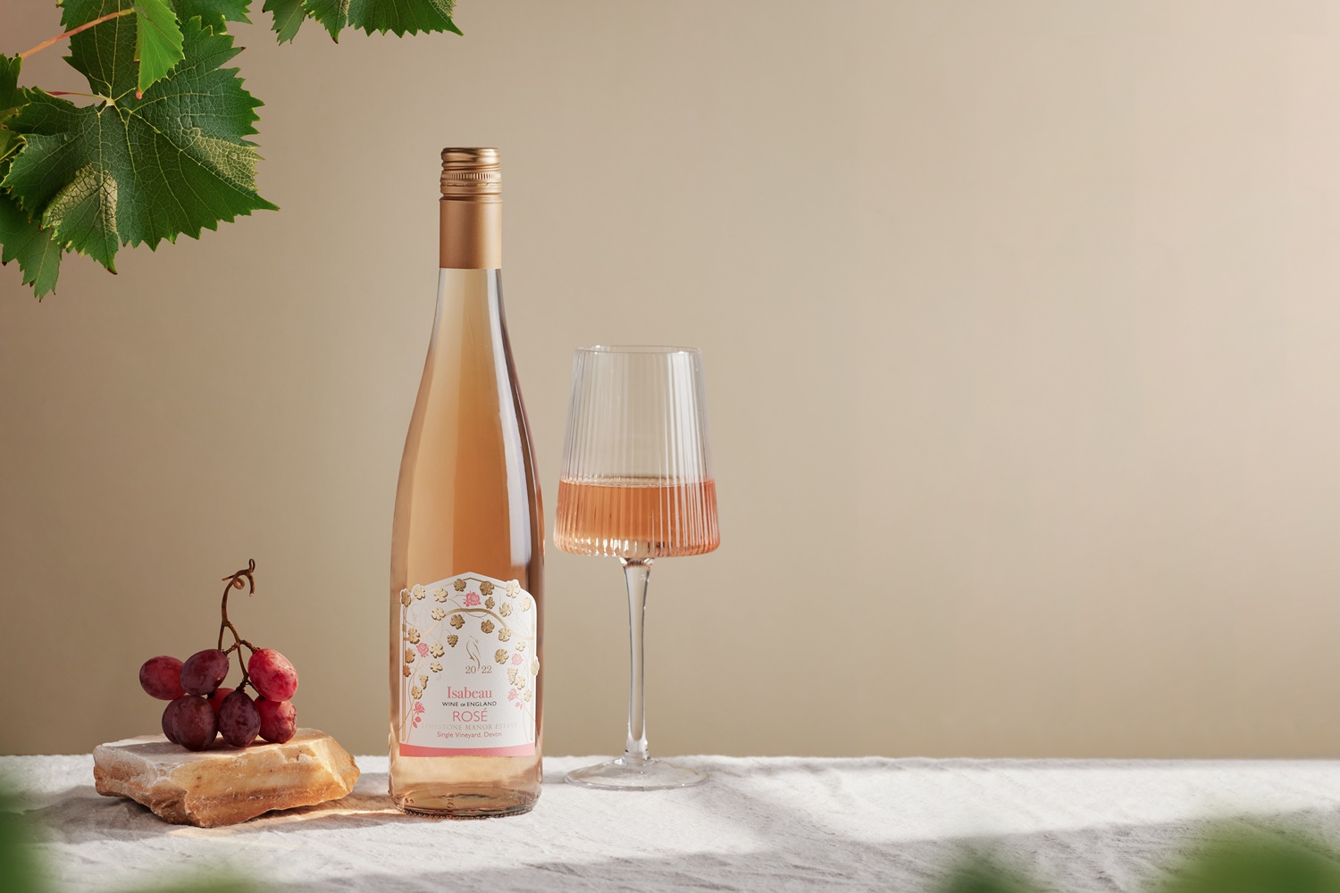 Lympstone Manor Isabeau Rose wine bottle displayed with a wine glass, grapes and vine leaves.
